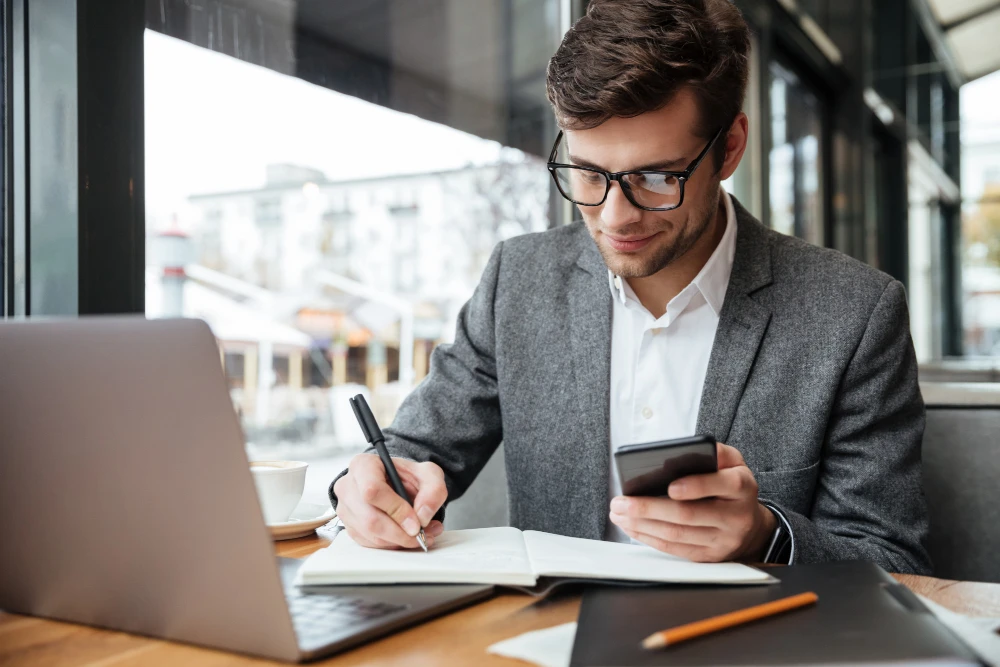 homme qui écrit sur un livre devant son ordinateur avec un téléphone à la main